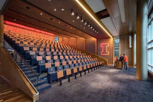 Auditorium with tiered seating, orange and blue University of Illinois branding. People near a podium.