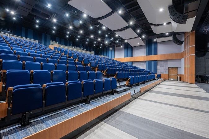 Auditorium interior with blue seats, wooden accents, and stage. Ceiling with lights and acoustic panels.