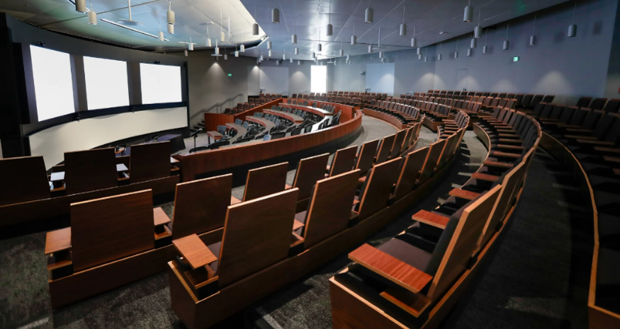 Curved auditorium with tiered seating, facing three large screens. Dark wood and dim lighting.