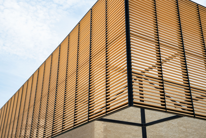 Corner of a building with a wooden slatted facade, tan against a light blue sky.