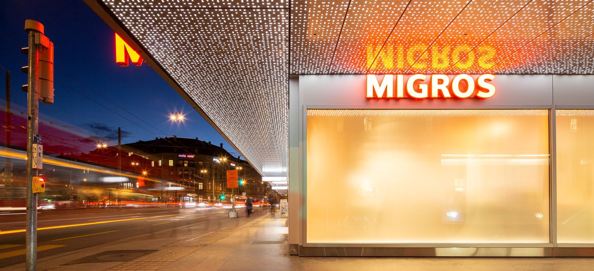 Nighttime street scene with a Migros store, neon sign, and long exposure light trails.