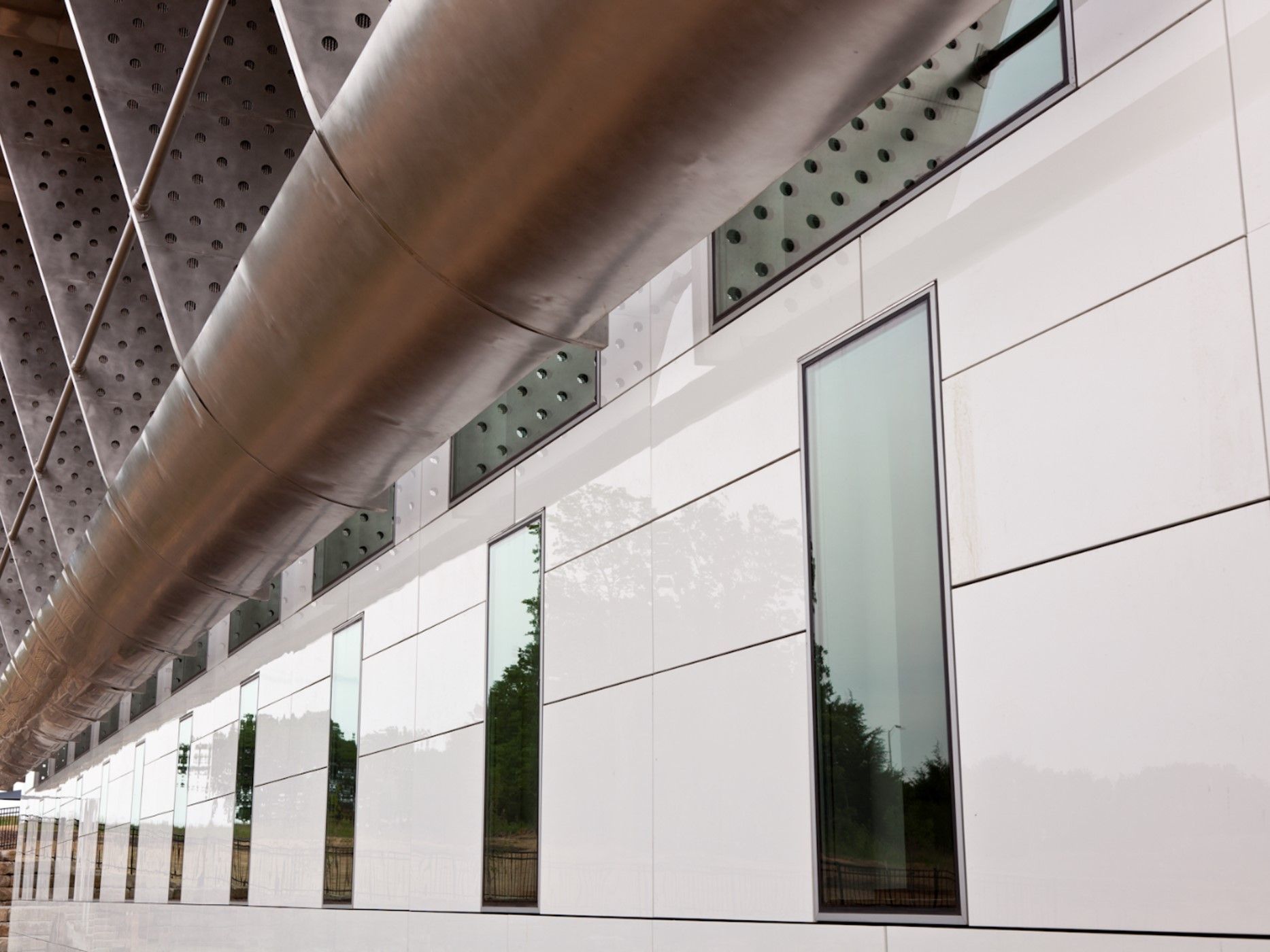 Modern building exterior with white panels, tall windows, and a metallic pipe running horizontally.