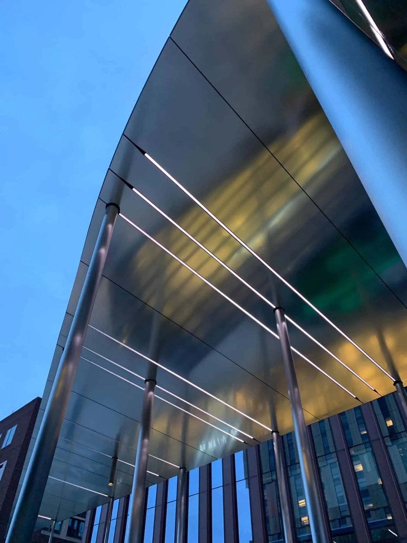 A modern building facade with a curved metal canopy supported by pillars, lit by white lights, and a blue sky background.