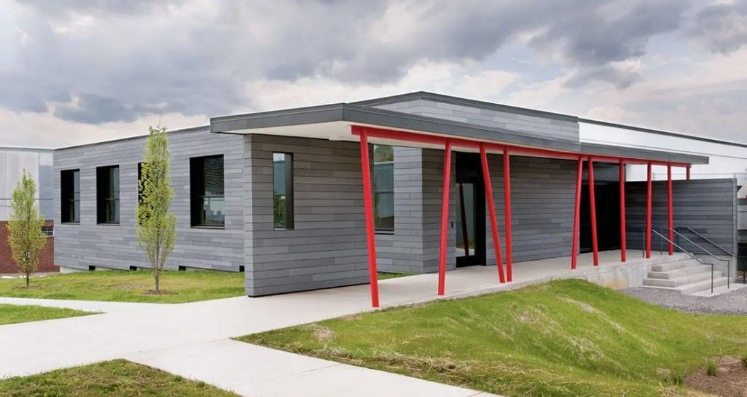 Modern gray building with red support beams, small trees, and a grassy lawn under a cloudy sky.