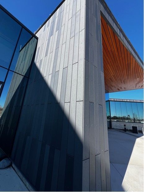 Modern building exterior with gray paneling, glass walls, and a wood-lined overhang under a blue sky.