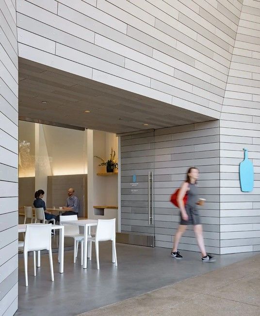 Person walking past an entrance with grey horizontal siding. Interior view includes tables, chairs, and other people.