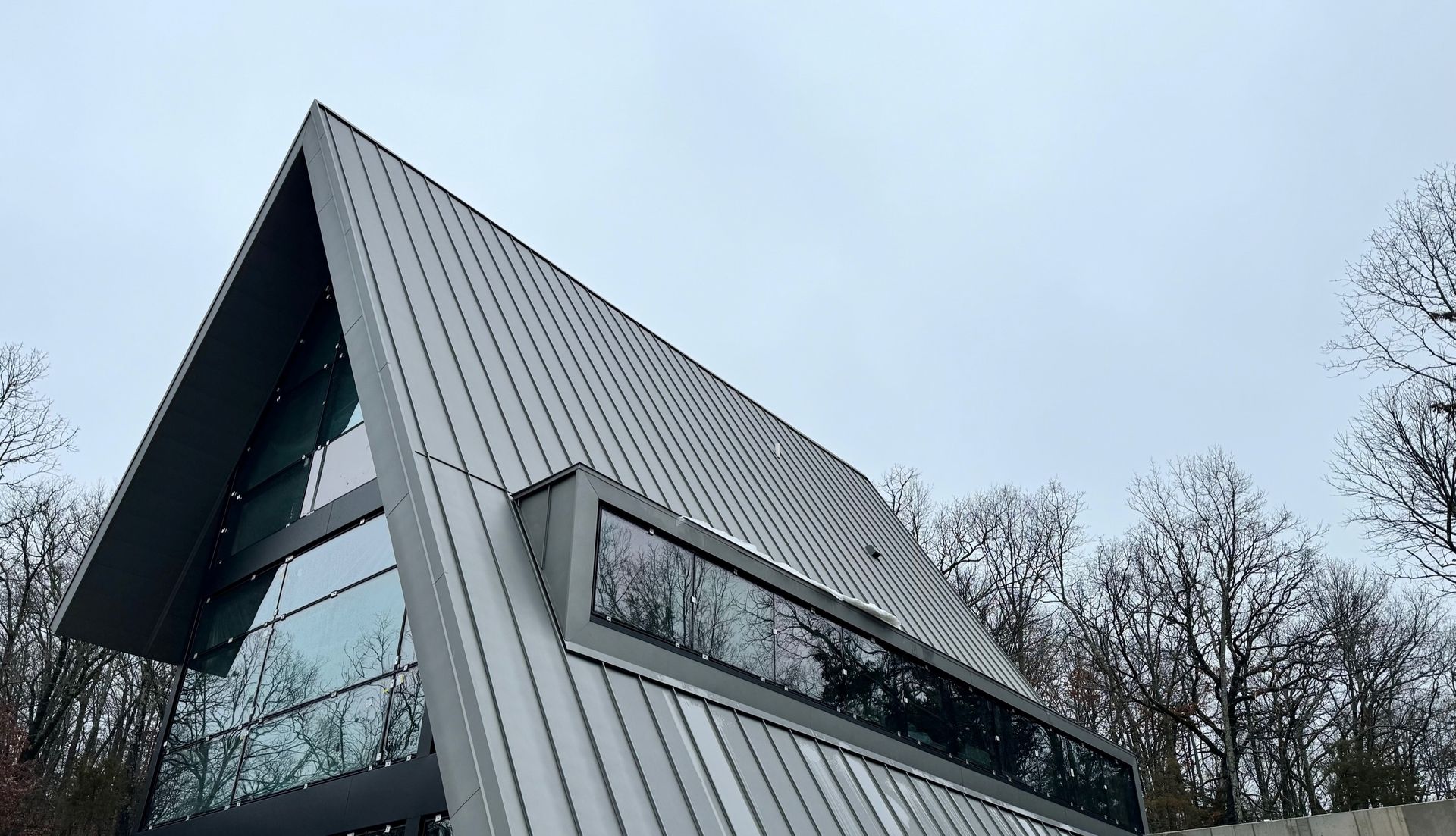 Modern building with slanted gray roof and large windows, against a cloudy sky and trees.