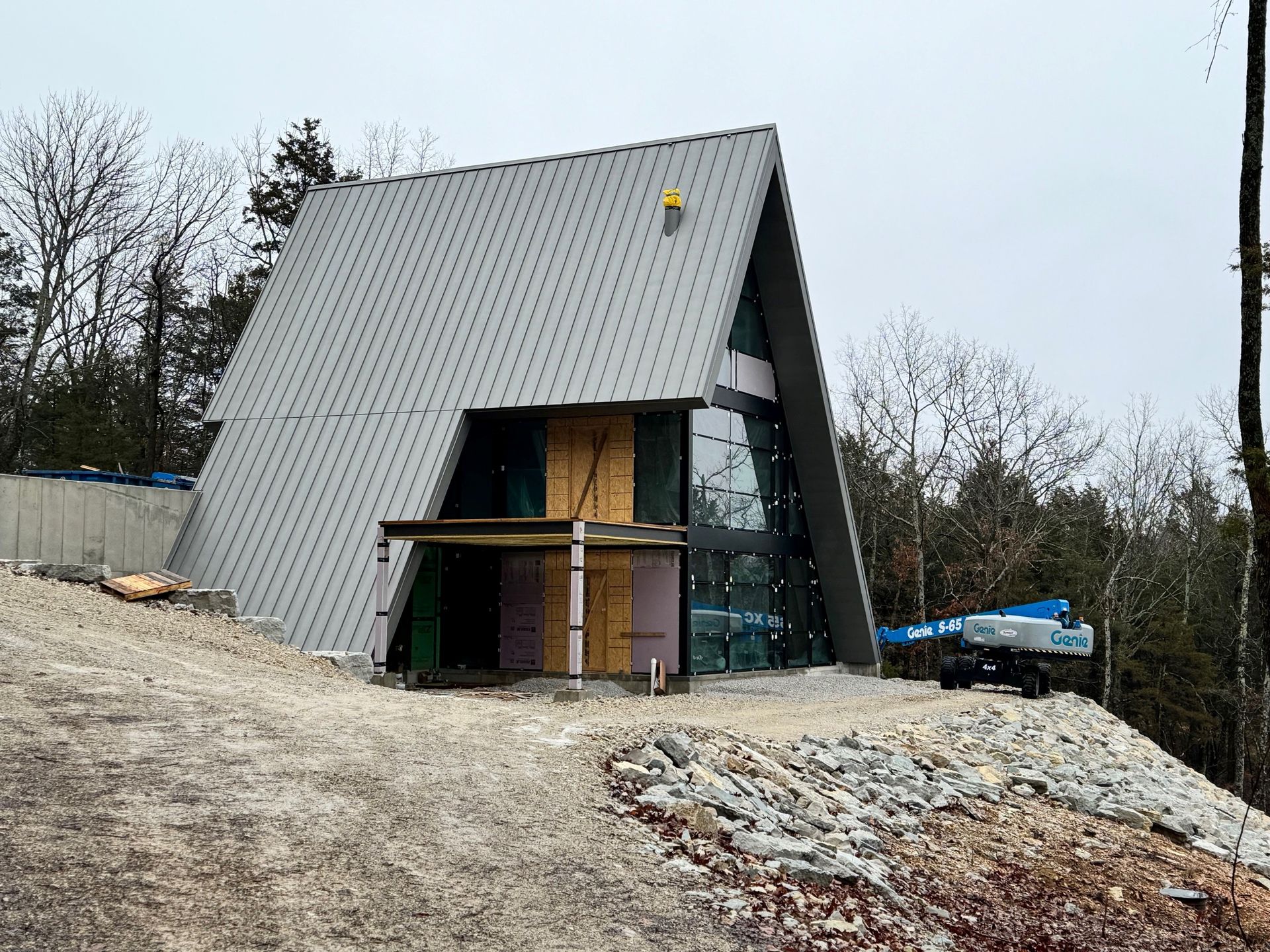 A-frame house under construction on a gravel lot; metal roof, large glass windows, and plywood accents.