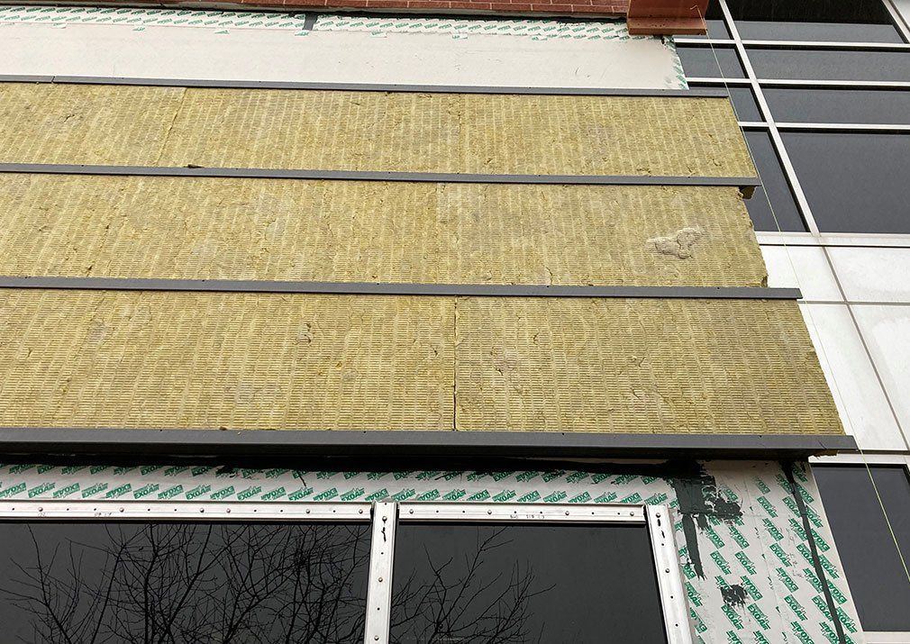 Exterior building wall showing installed insulation panels between horizontal metal strips, next to windows.
