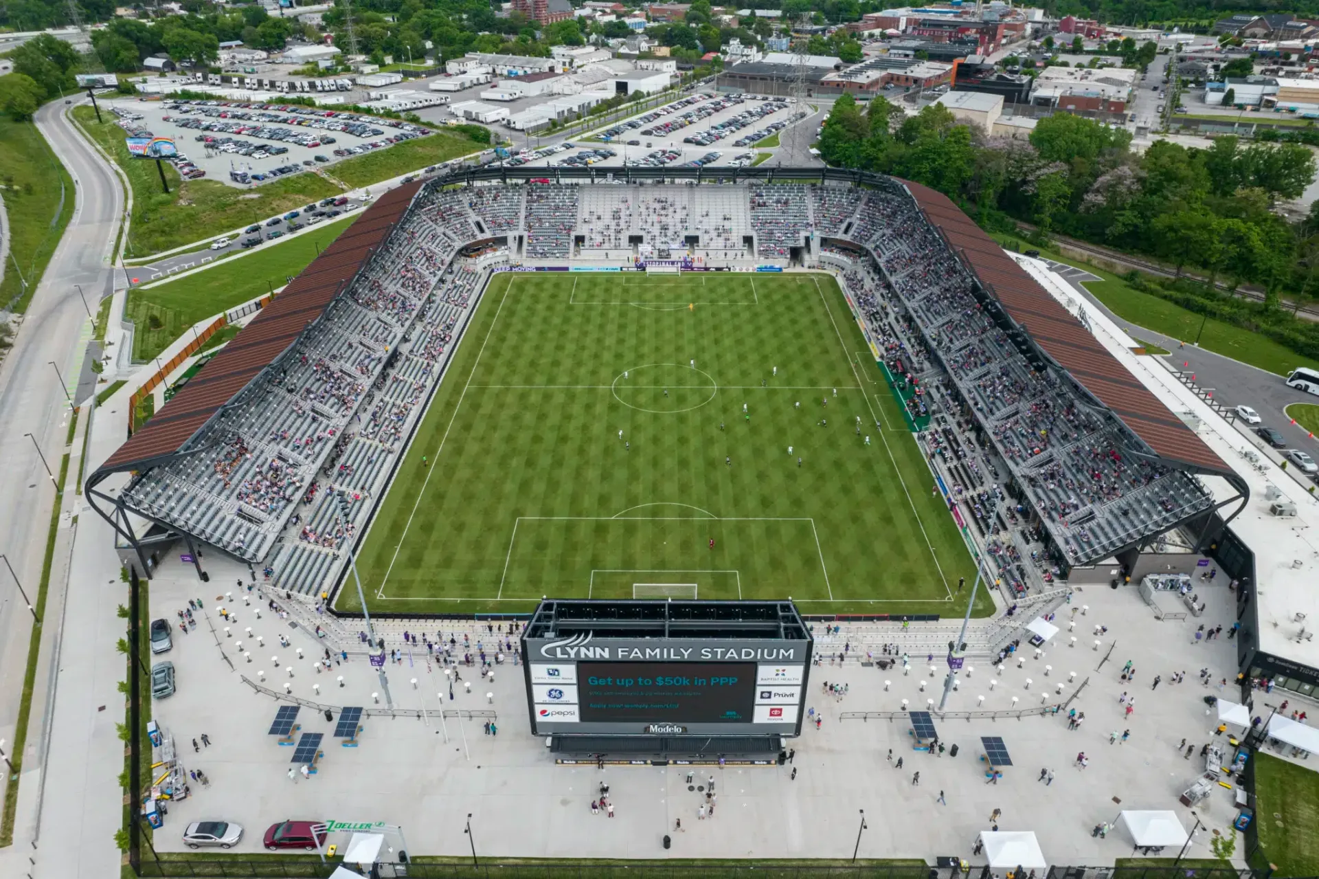 Aerial view of a packed soccer stadium with a green field and large scoreboard, with a city in the background.