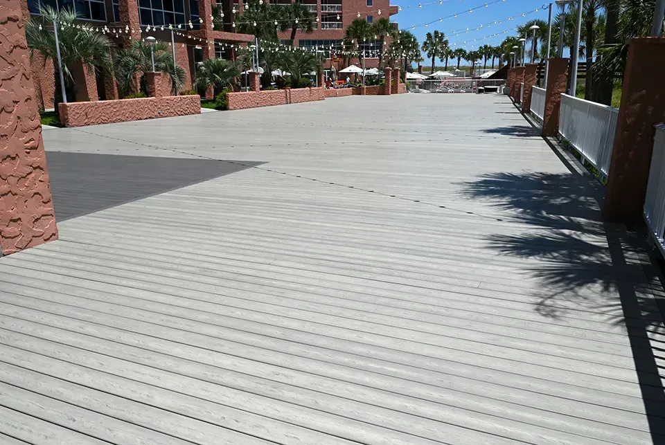 Wide gray wooden walkway leading toward a resort with palm trees. Sunny day, shadows cast.