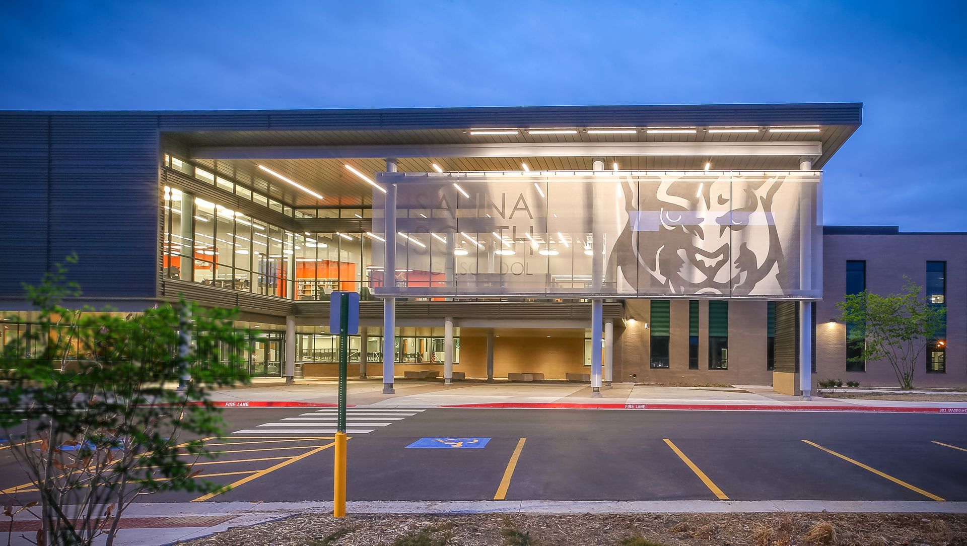 Modern school building exterior with illuminated lion logo at entrance, dusk. Gray, orange, and glass details.