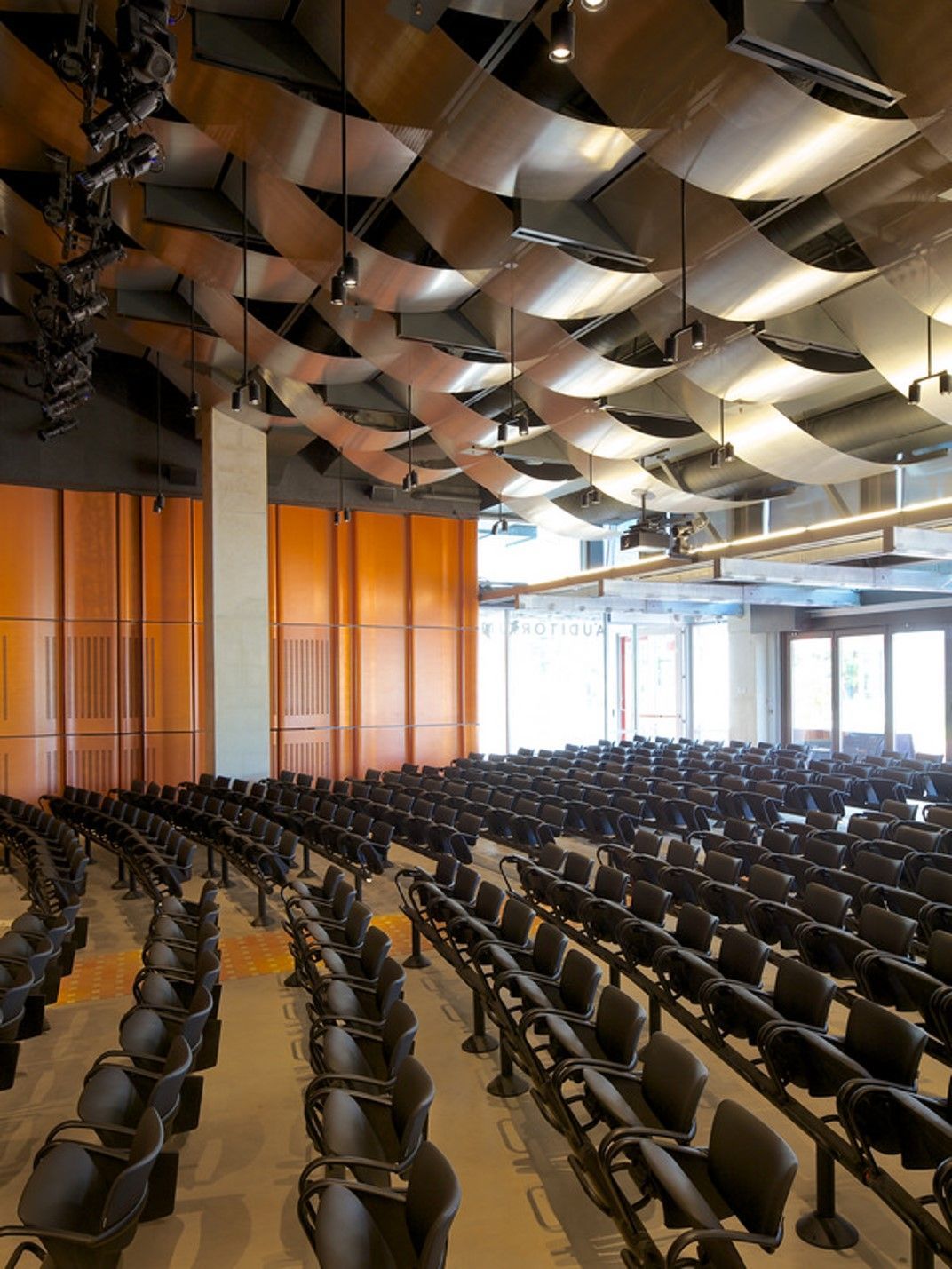 Auditorium with rows of seats facing a stage. Metal ceiling panels hang above.