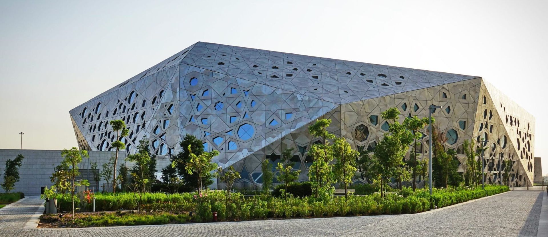 Modern building with irregular facade, dotted with various sized openings, set against a blue sky, surrounded by greenery.