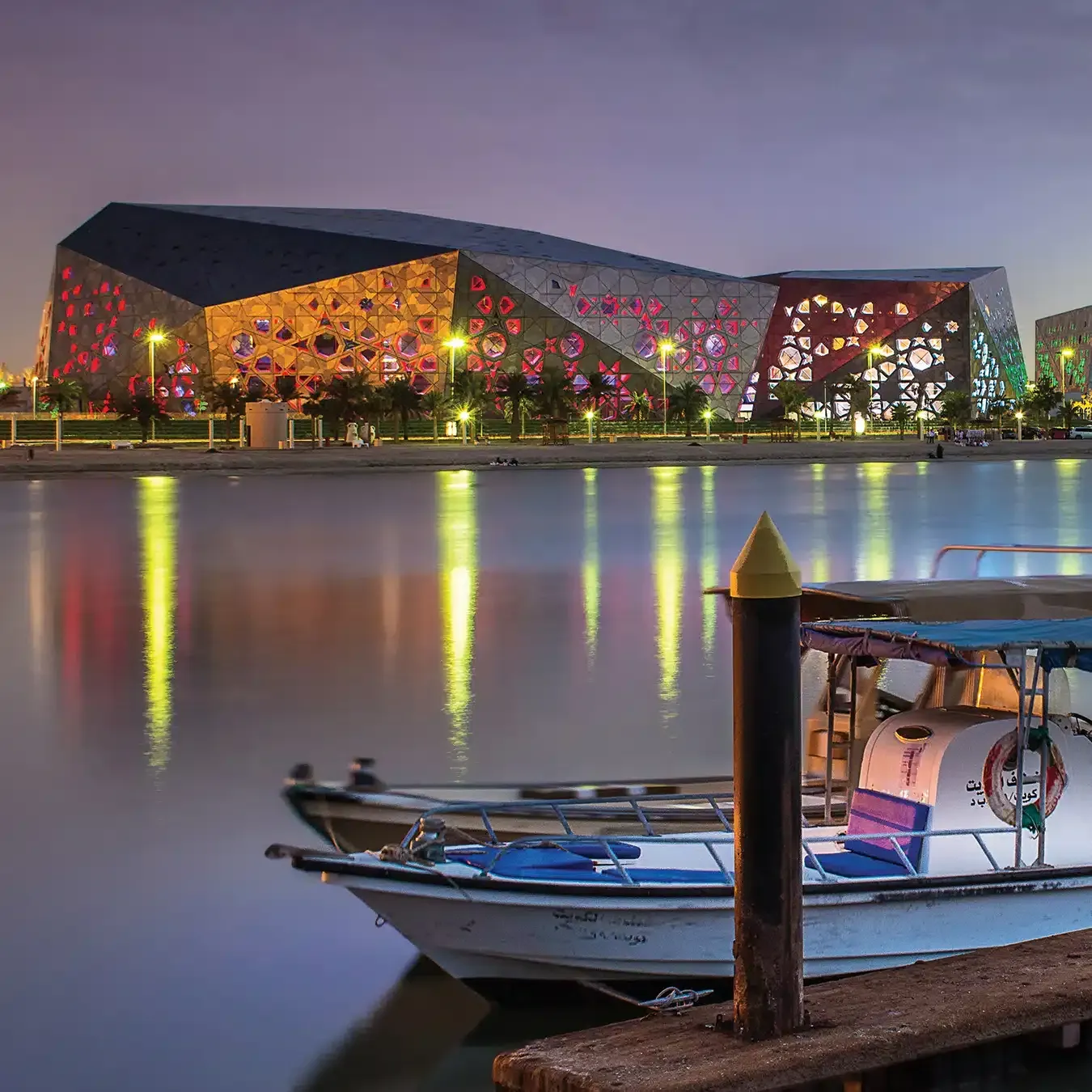 Large modern building with geometric facade, lit up at night, reflected in calm water. Small boats in the foreground.