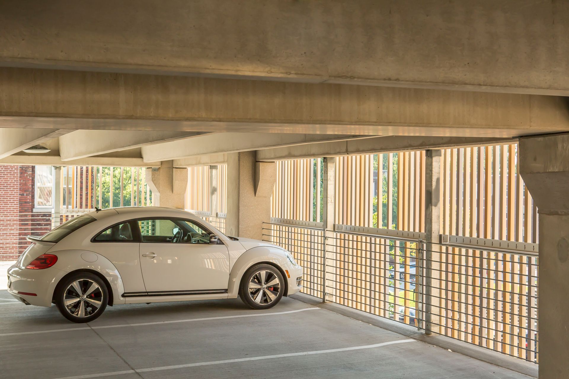 White Volkswagen Beetle parked in a concrete parking garage, next to a vertical slatted barrier.