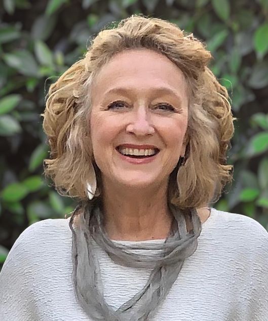 Woman with blonde curly hair, smiling, wearing a light-colored top and gray scarf; outdoor setting.