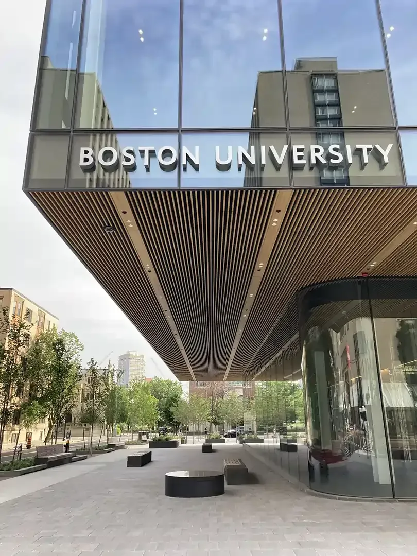 Boston University building exterior with large glass facade; benches and trees in foreground.