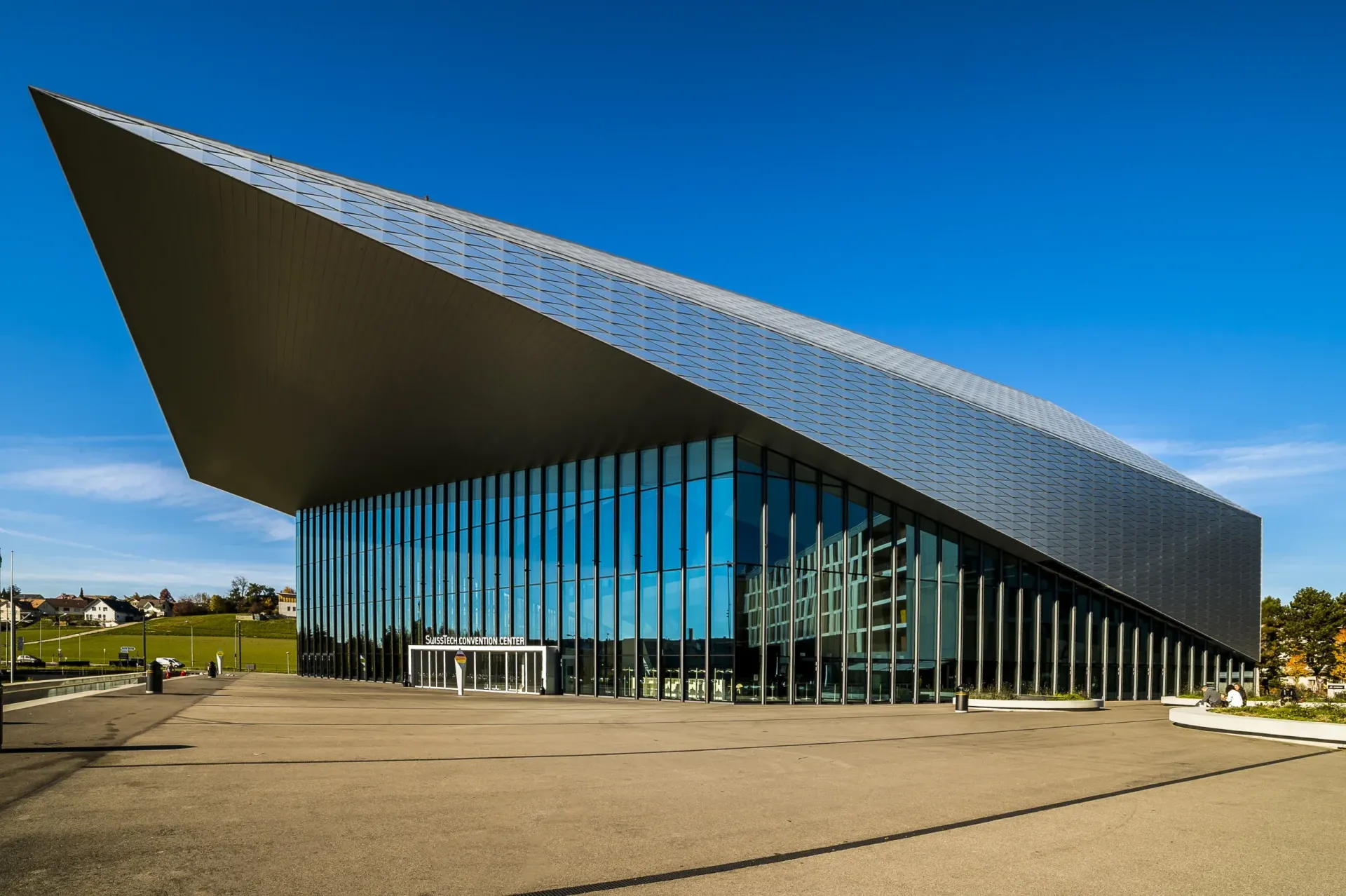 Modern building with angled metallic roof, glass walls, and bright blue sky.