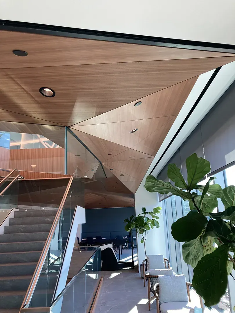 Geometric wood ceiling above a staircase with glass railing. A potted plant sits near a window.