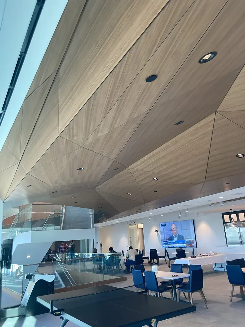 Modern interior with wood paneled ceiling, blue chairs, tables, and a screen showing a person.