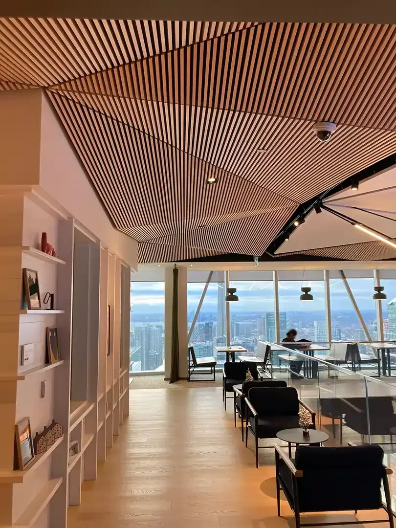 Modern office interior with wooden slat ceiling, floor-to-ceiling windows overlooking cityscape, lounge seating.
