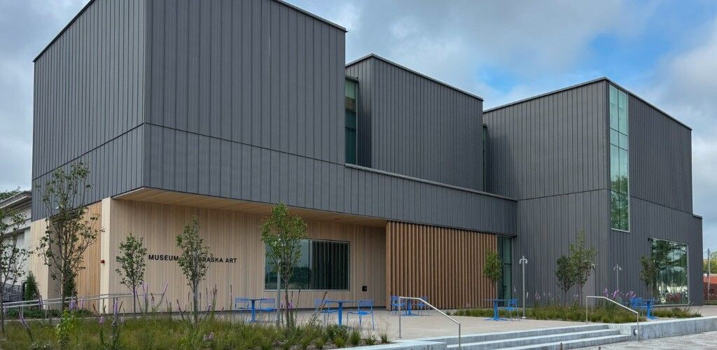 Modern building with gray metal siding and light wood paneling, fronted by landscaping and a blue sky.