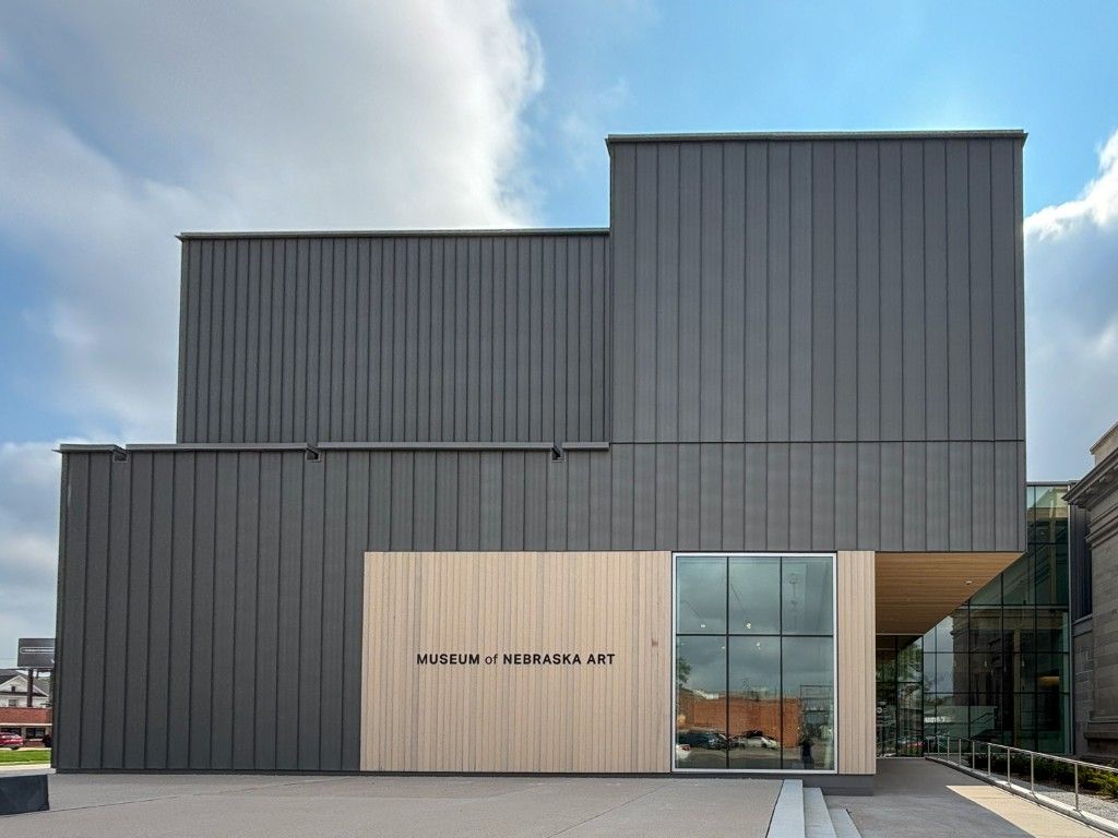 Gray, modern building with a light wood entrance, glass facade, and a blue sky.