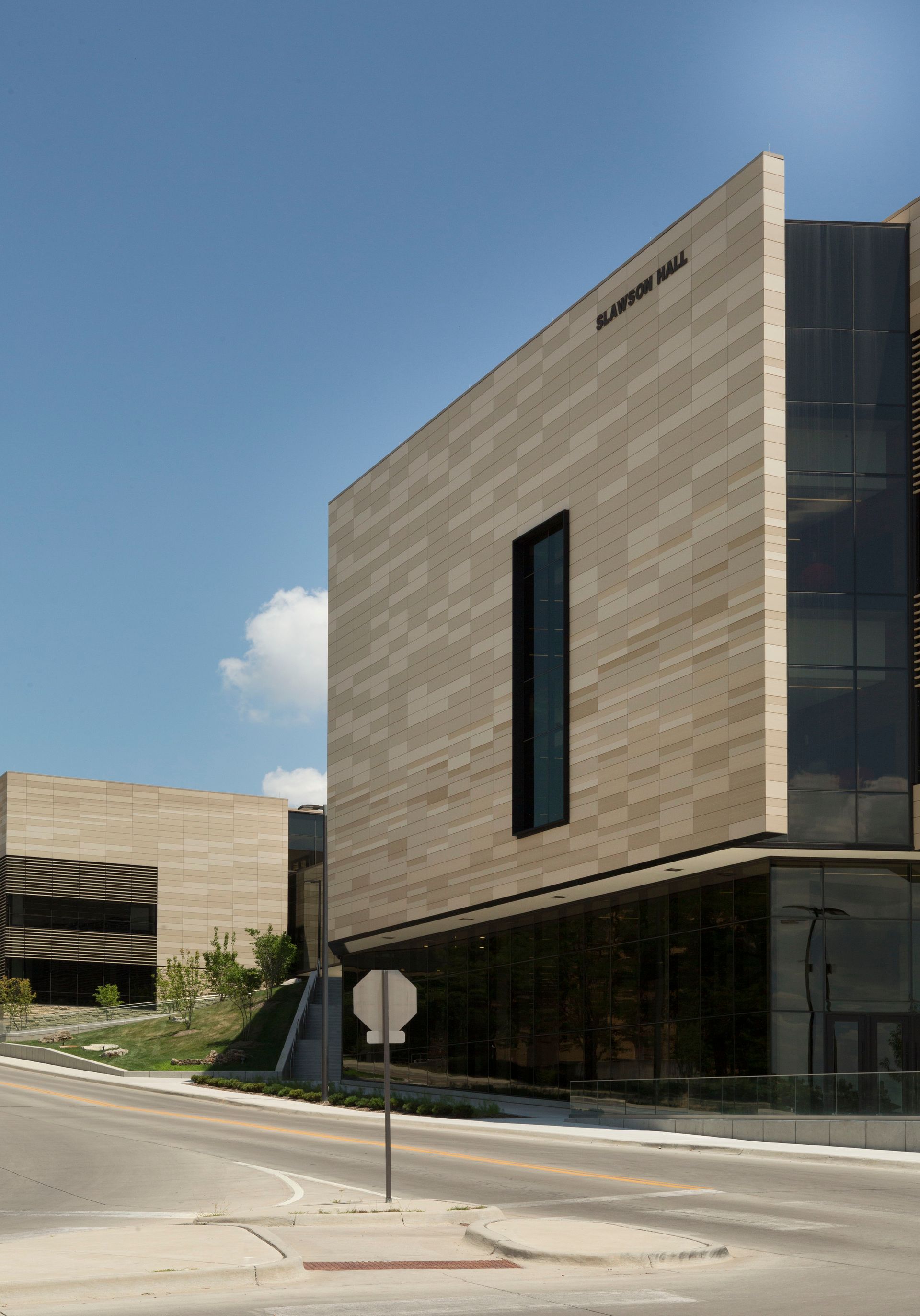 Modern tan stone building with a rectangular window, blue sky, and a street.