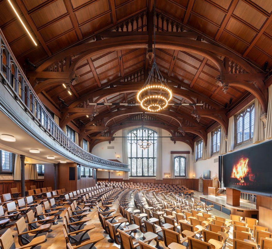 Ornate wooden lecture hall with curved rows of seats facing a screen and stage. A balcony overhangs the left side.