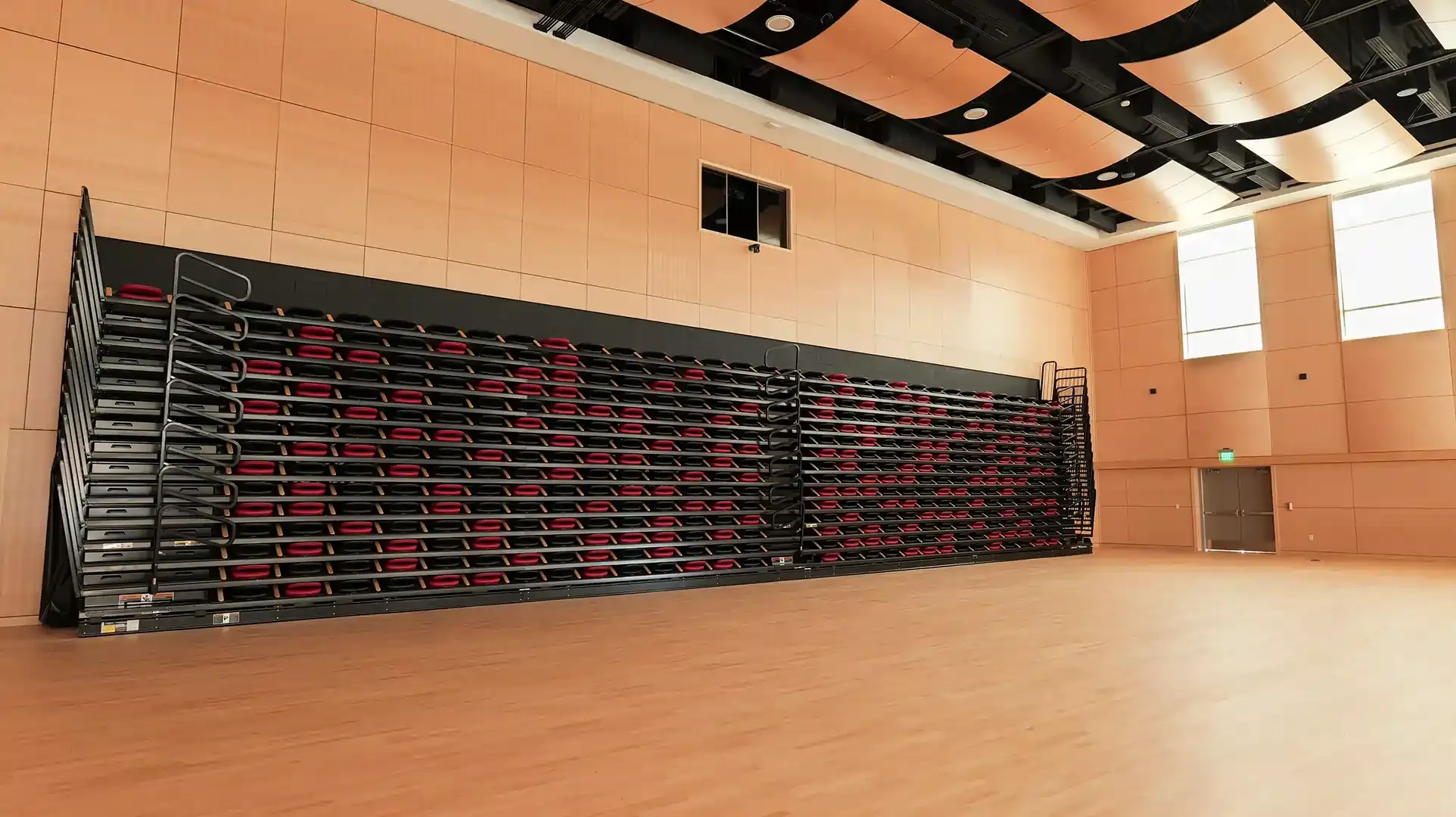 Large, open room with retractable seating folded against a wall. Wooden floor and walls, with windows and a door visible.