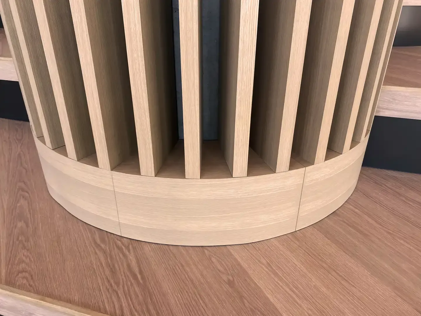 Wooden, cylindrical architectural element with vertical slats, set on wood flooring.