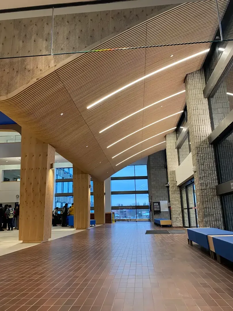 Wooden ceiling with curved beams and recessed lights in a modern building with large windows and stone columns.