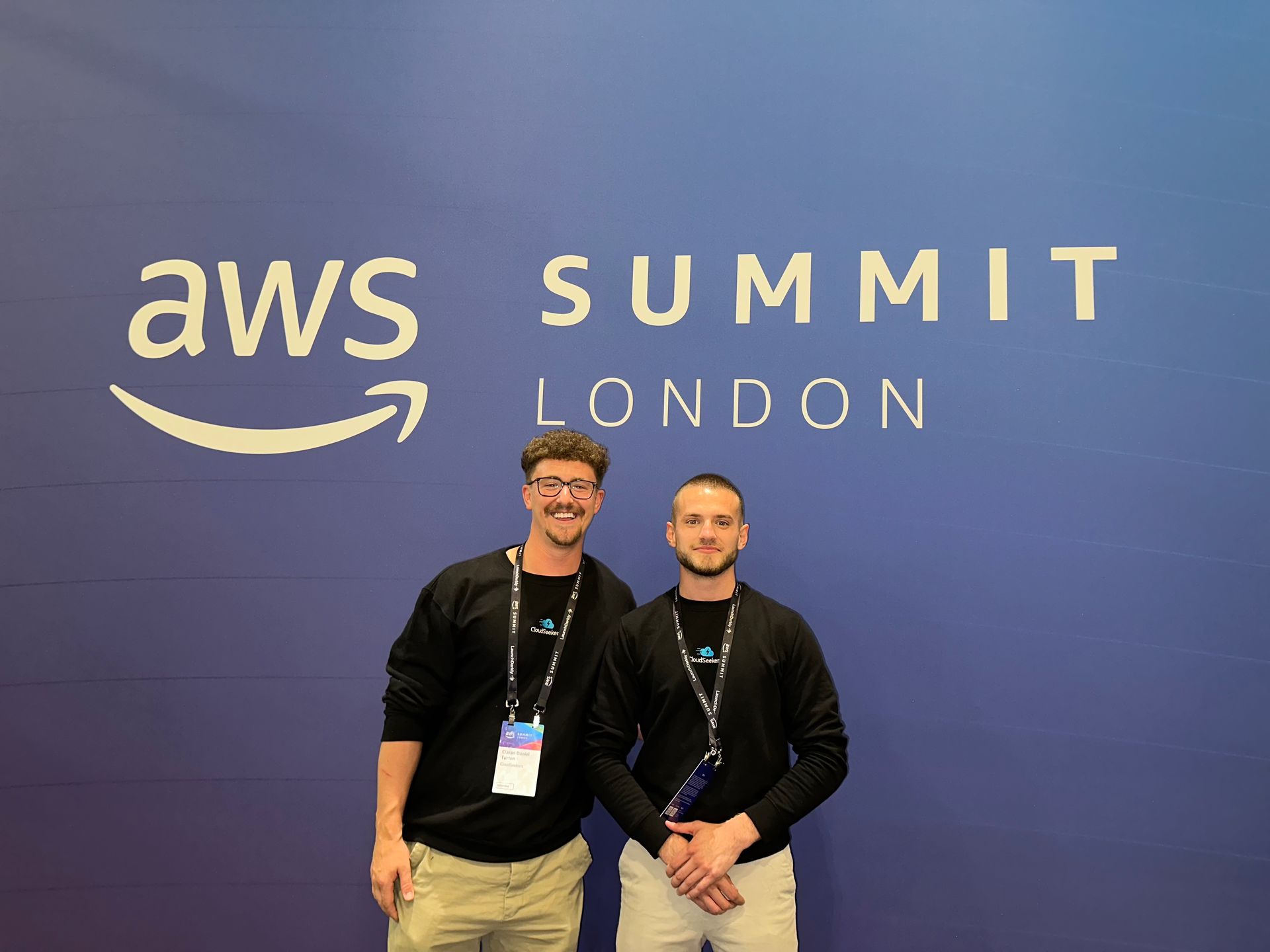 Two smiling people wearing lanyards stand in front of a blue AWS Summit London backdrop.