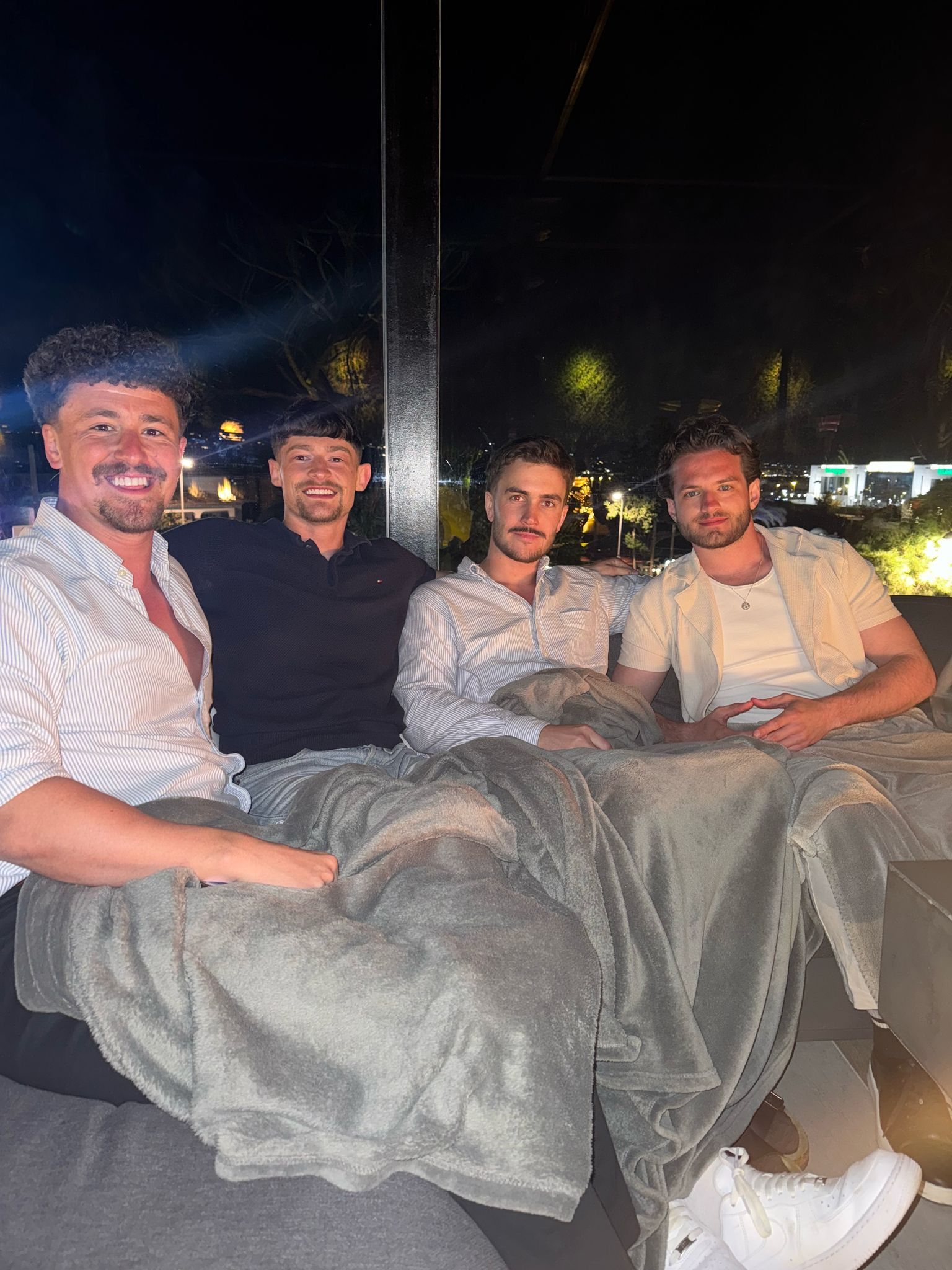 Four men sit together outdoors at night, wrapped in a large, shared gray blanket, smiling toward the camera.