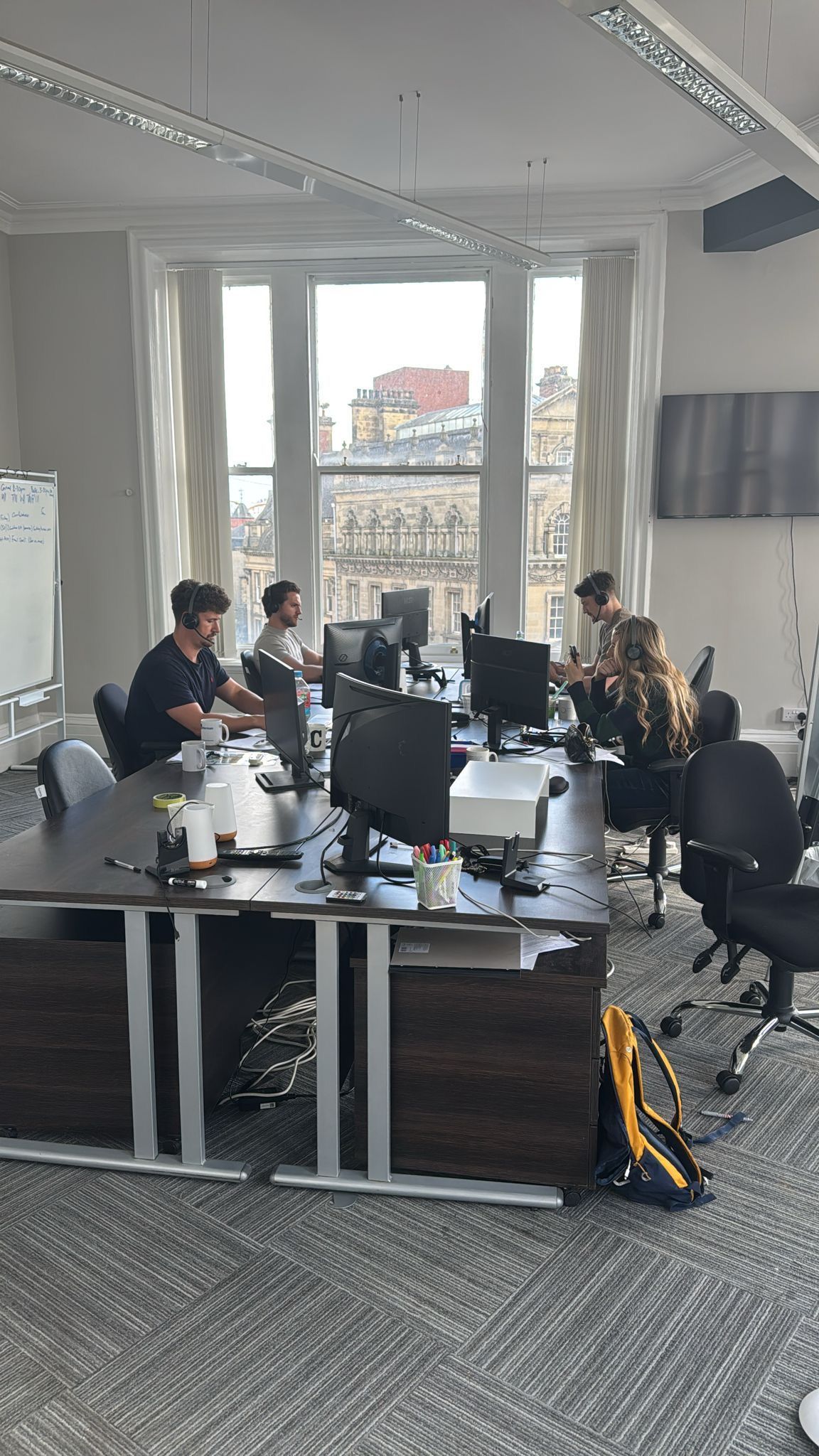 Four people working at desks in a bright office with large windows overlooking a city street.