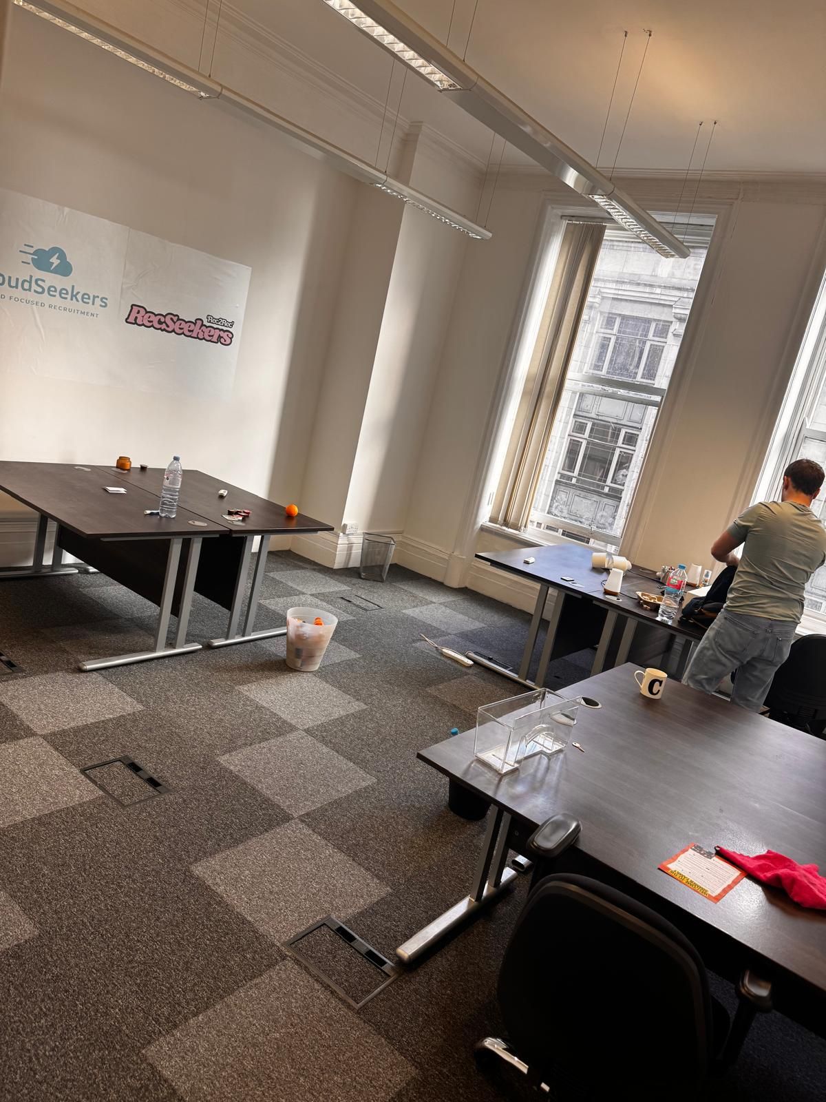 A bright, modern office with tables arranged in an open space. One person stands by a large window overlooking a street.