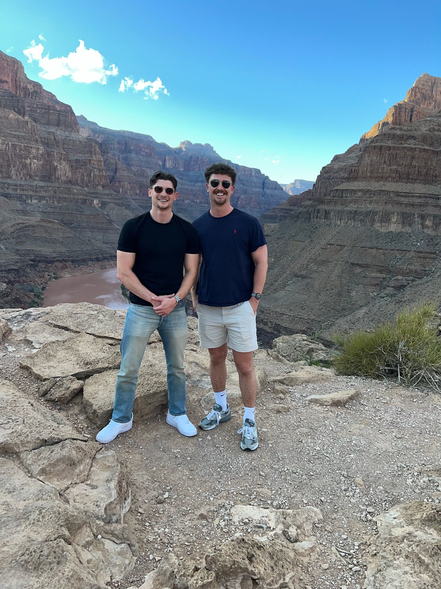 Two men wearing sunglasses stand side-by-side on a rocky overlook with a canyon and river in the background.