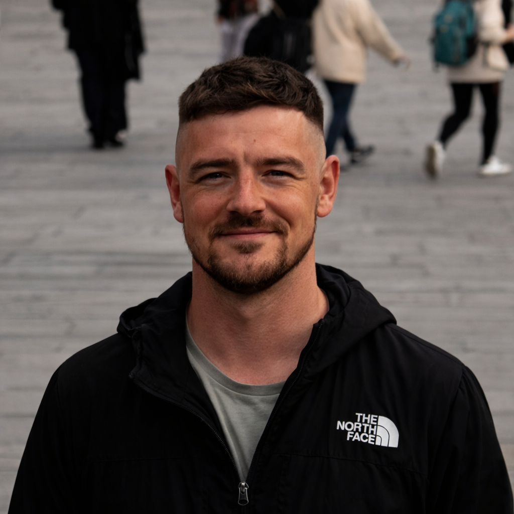 A person with short, dark hair and a beard, wearing a black North Face jacket, smiling outdoors with pedestrians behind.