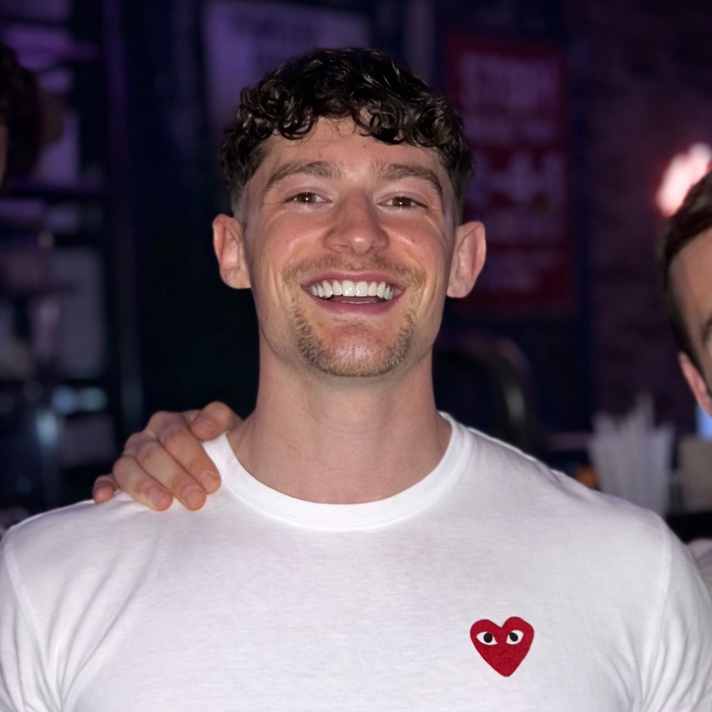 A smiling person with curly brown hair wearing a white t-shirt with a small red heart logo, indoors at night.
