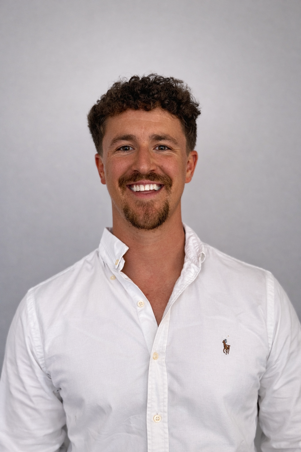 A person with curly hair and facial hair, smiling in a white button-up shirt against a light grey background.