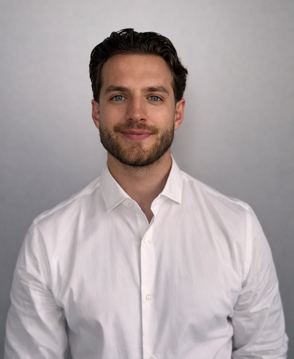 A person with dark hair and a beard, wearing a white button-down shirt, smiles against a plain gray background.
