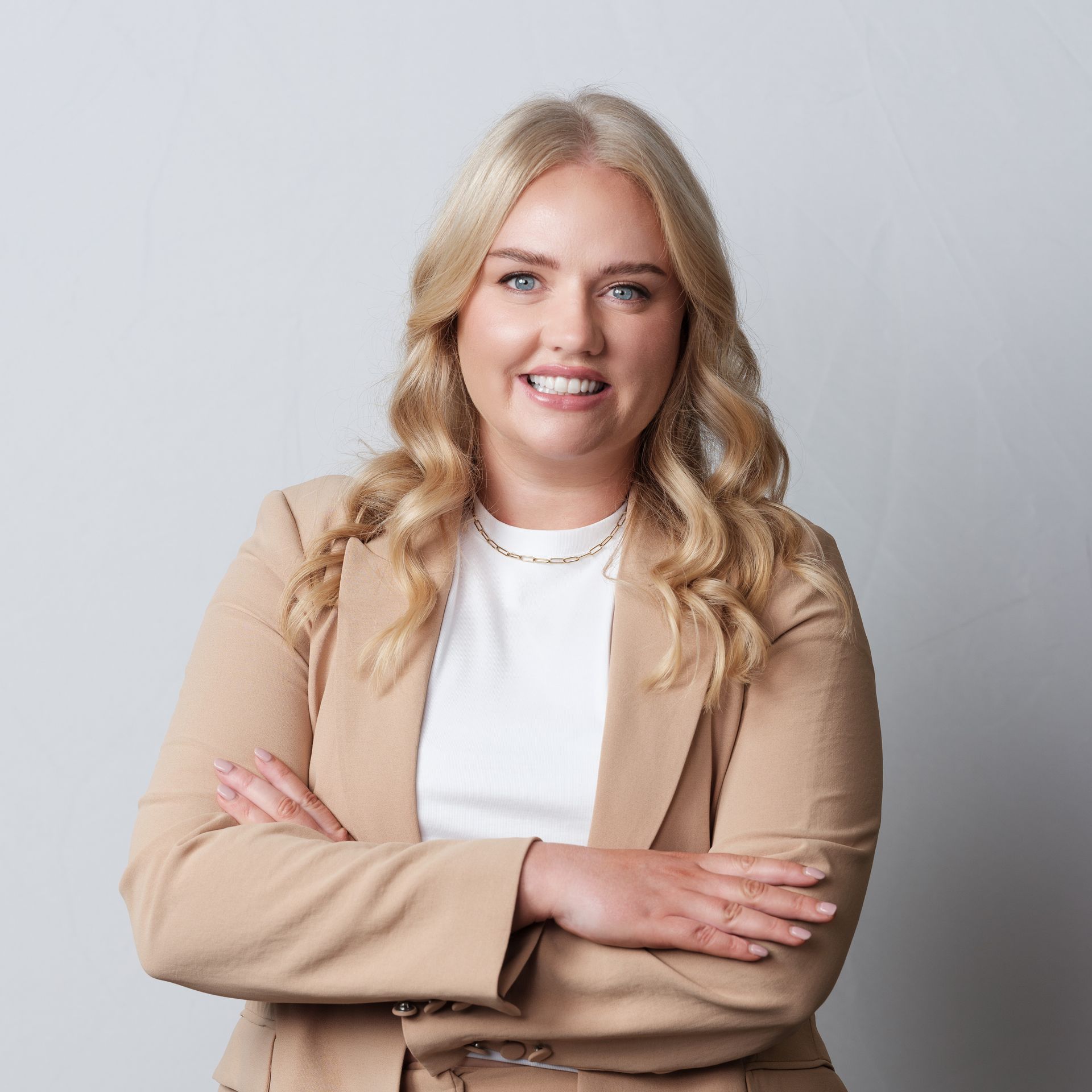 Woman with blonde hair, wearing a tan blazer, arms crossed, smiling, against a gray background.