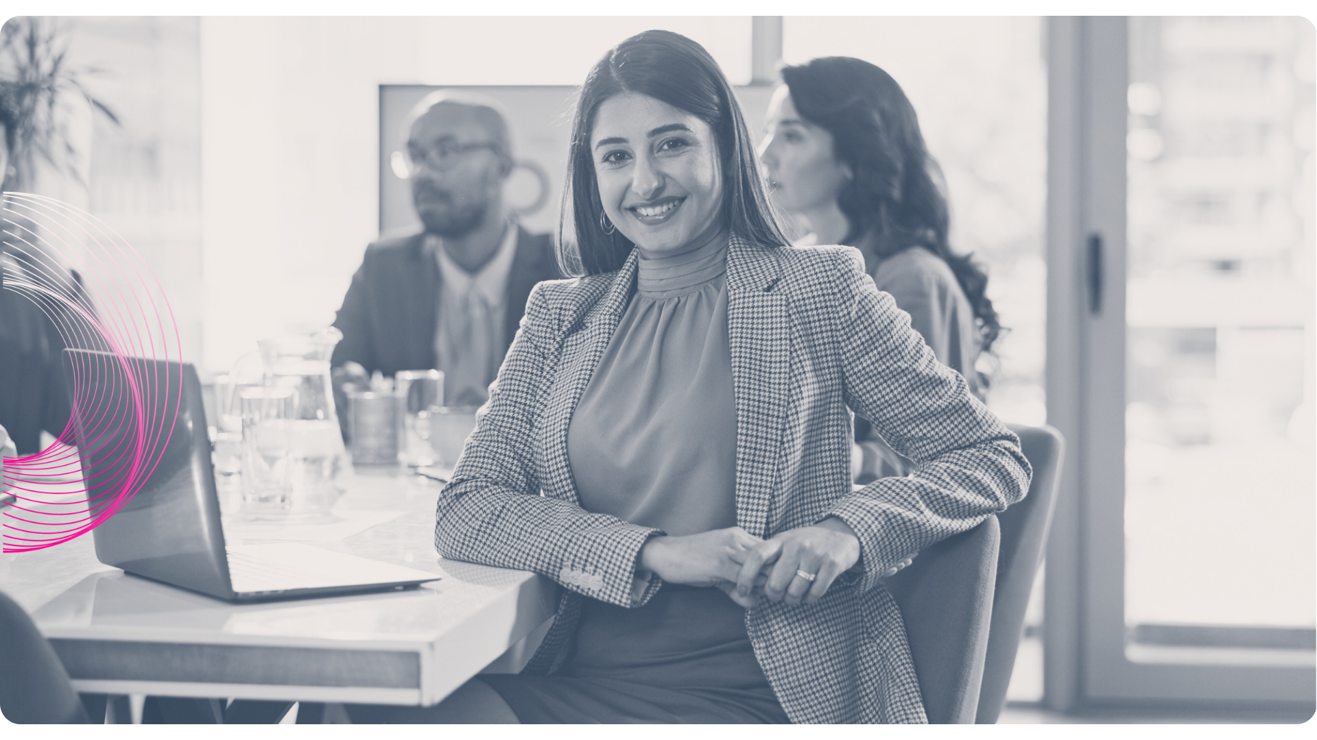 Woman smiles at the camera, seated at a table in an office, with coworkers in the background.