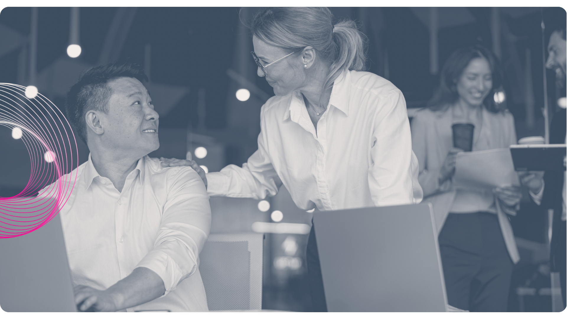 People in office setting working on laptops, one woman smiling at a man, others in background.