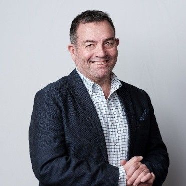 Man in blazer and checkered shirt smiles, hands clasped, against a light gray background.