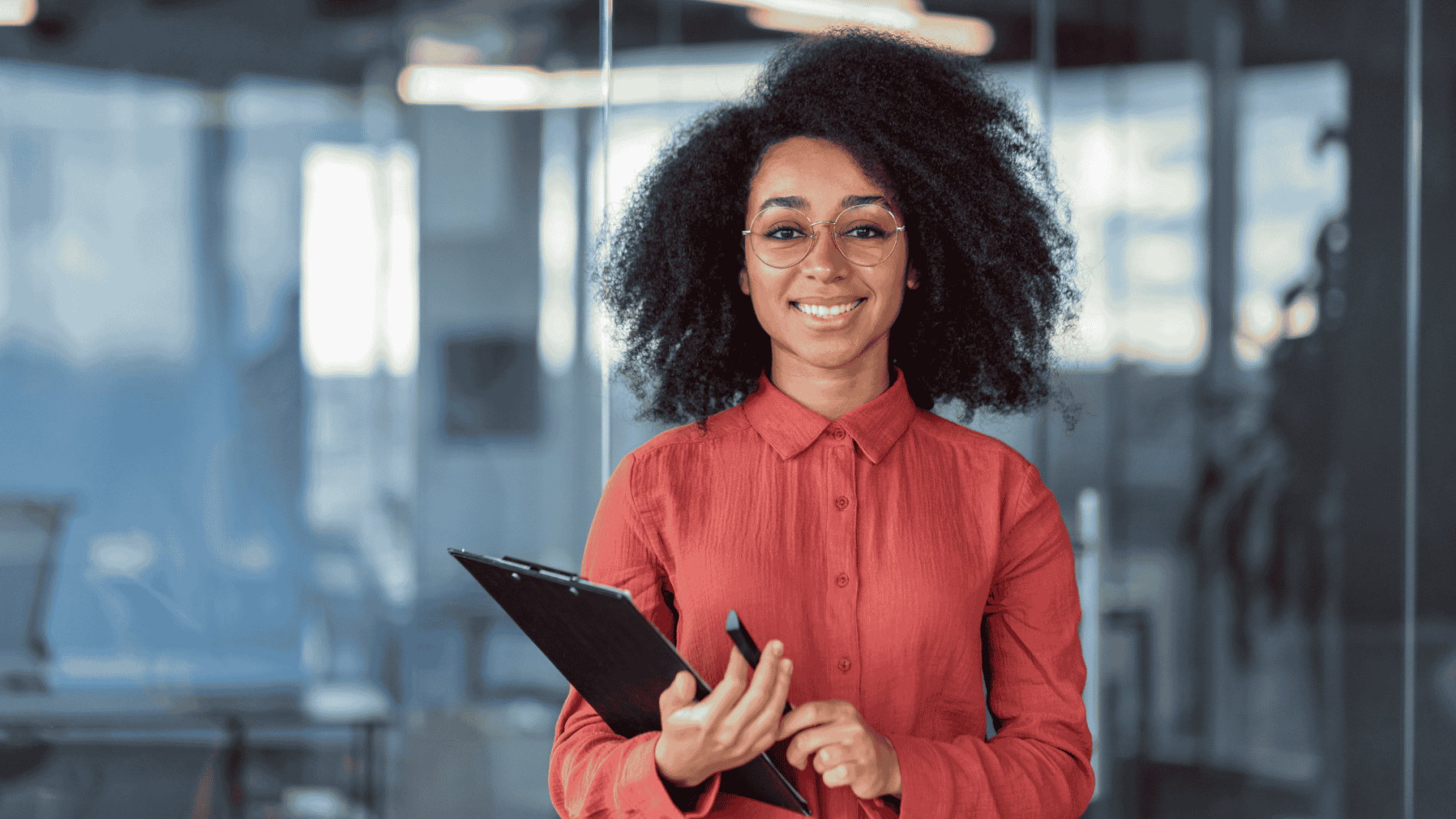 Woman with glasses and curly hair smiles, holding a clipboard in a modern office setting. She wears a red shirt.
