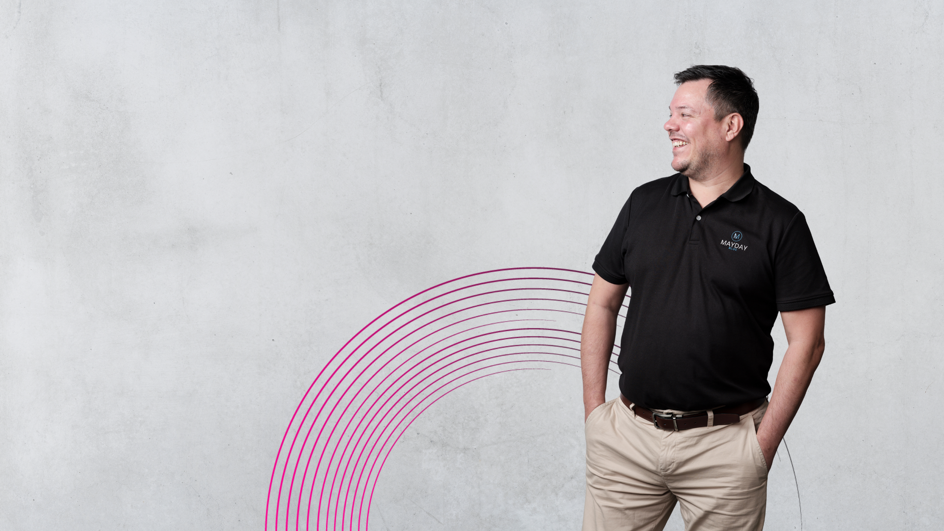 Man in black polo shirt and khaki pants, smiling, hands in pockets, against a gray wall with pink arcs.