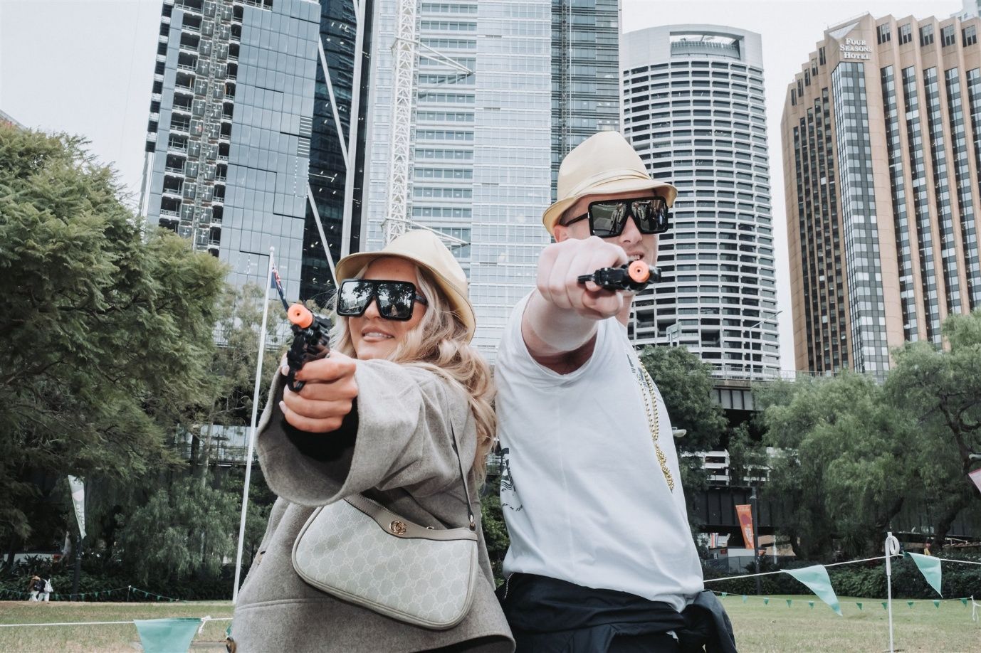 Two people, wearing hats and sunglasses, point toy guns at the viewer in a park, city buildings in background.