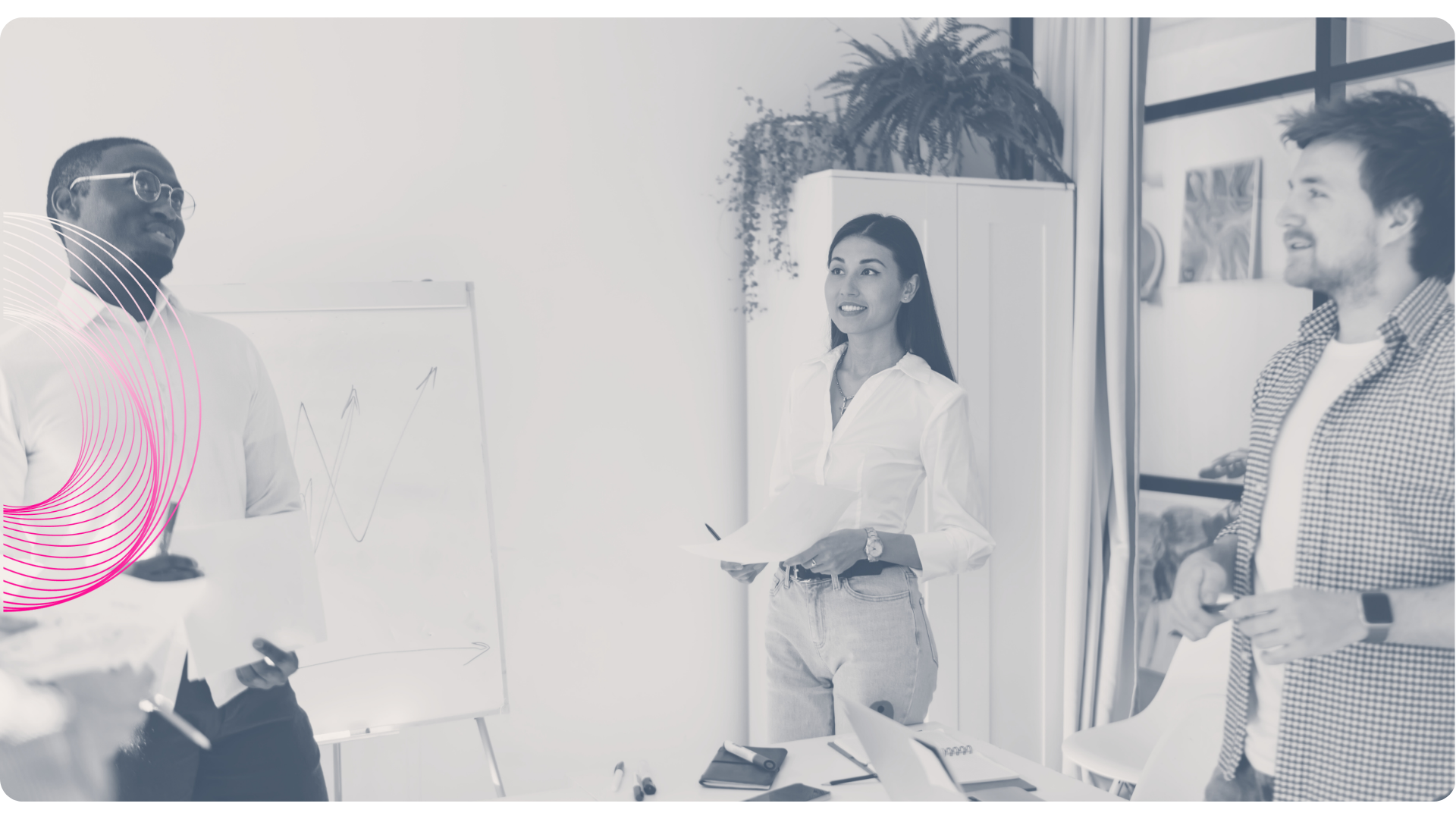 Three people in a meeting, one speaking at a whiteboard, the others listening, in an office.